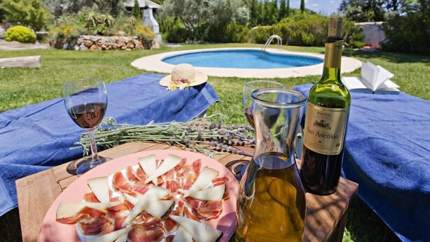 A poolside setting with a table set for a meal, including a plate of prosciutto, a bottle of wine, and glasses. - Villa Margarita Rocabella (El Chorro)
