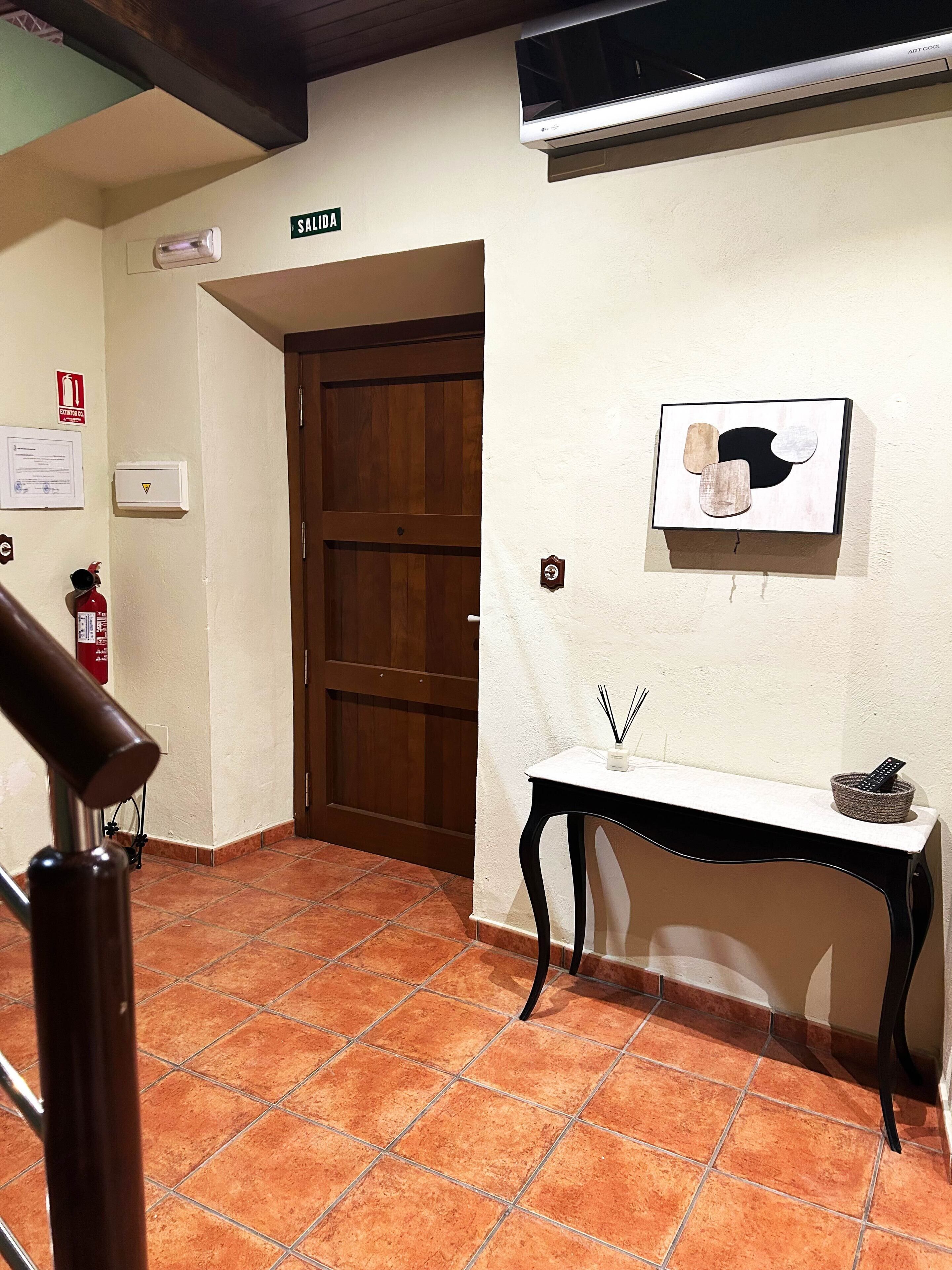 Un corridoio con una porta in legno, un muro bianco con un quadro incorniciato, un tavolino con un vaso e un telecomando, e un estintore.