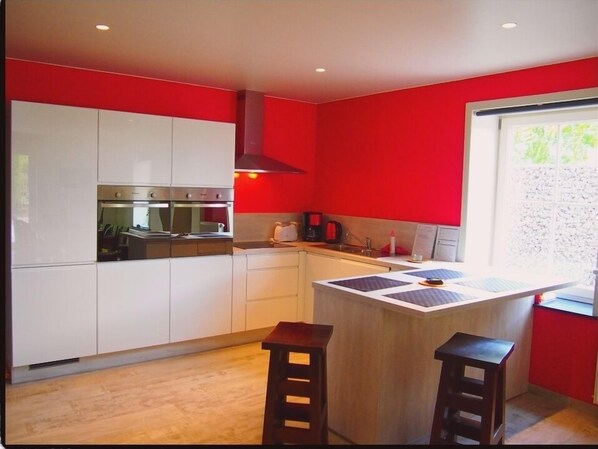 Een moderne keuken met witte kastjes, een inbouwoven en een rode accentmuur. Twee krukken en een raam met uitzicht op groen.