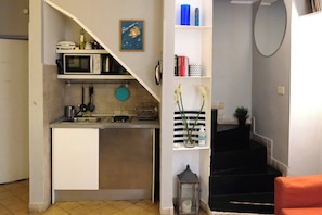 Uma cozinha compacta com micro-ondas, pia e um pequeno balcão. Há uma escada que leva a um patamar com um espelho e uma prateleira com livros e objetos decorativos.