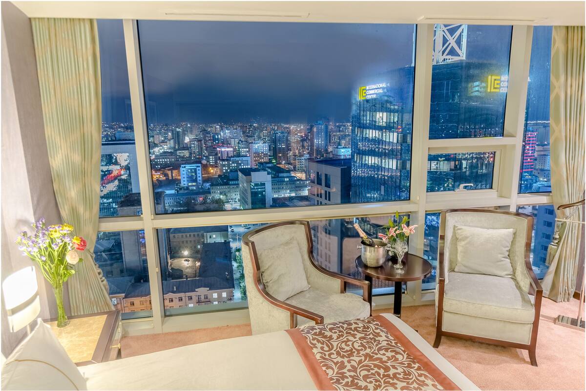 Habitación de hotel con un ventanal grande que ofrece vistas de la ciudad por la noche. Hay dos sillones, una mesita con un jarrón de flores y una cama con colcha estampada.