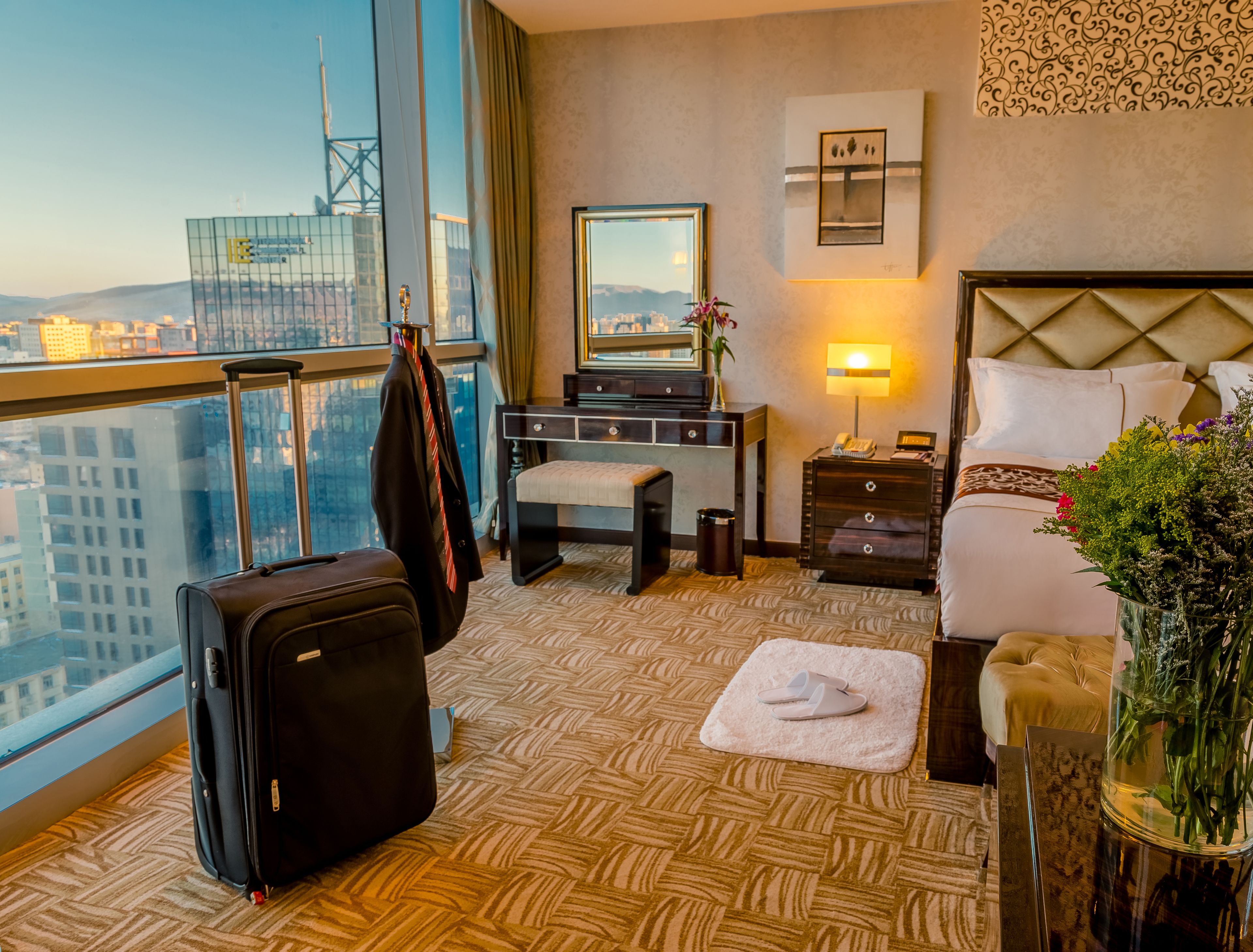 A hotel room with a large window, a desk, a bed, a suitcase, and a vase with flowers.