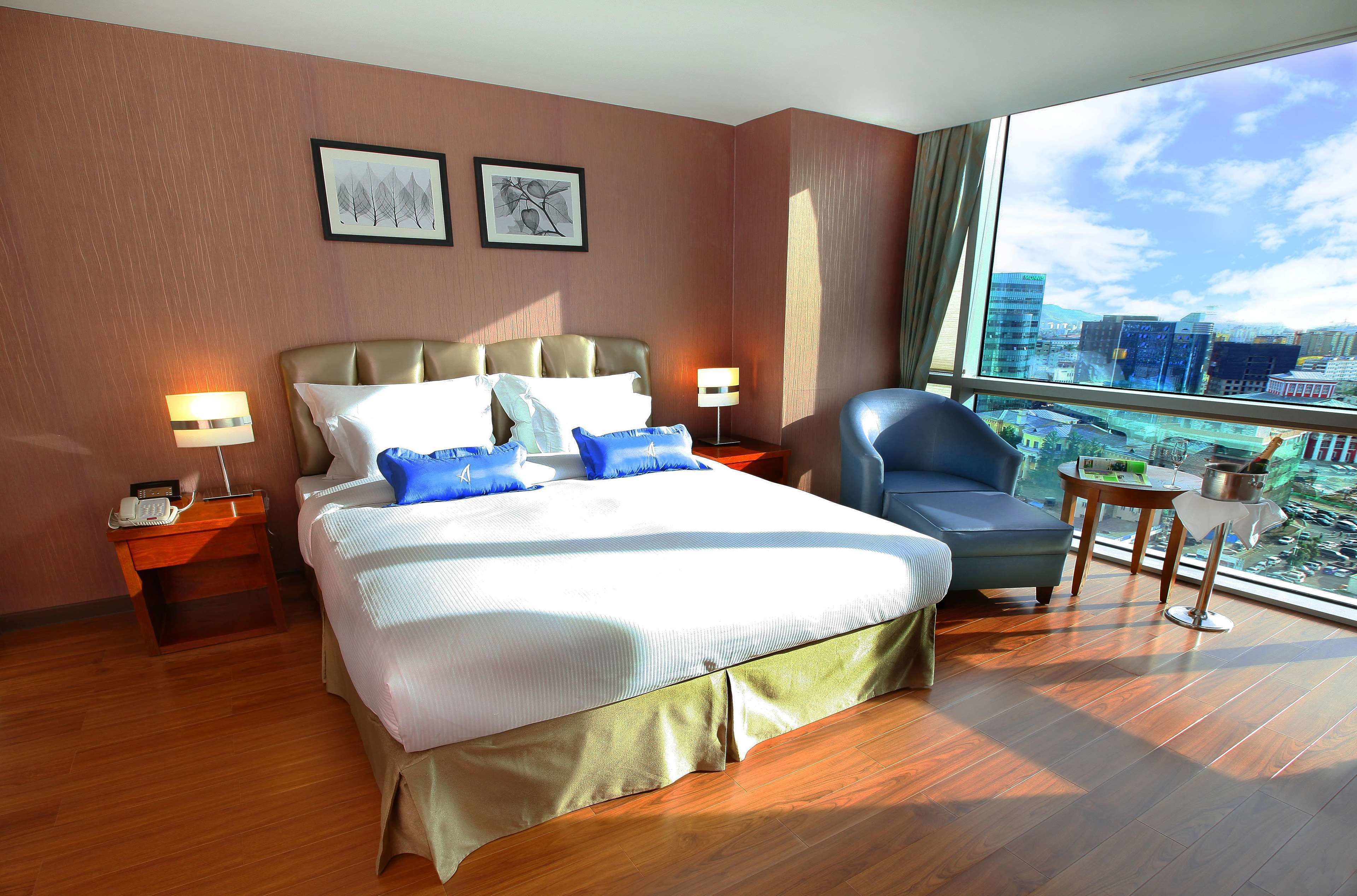 Habitación de hotel con una cama grande, una silla, una mesita y vistas a la ciudad a través de amplios ventanales.