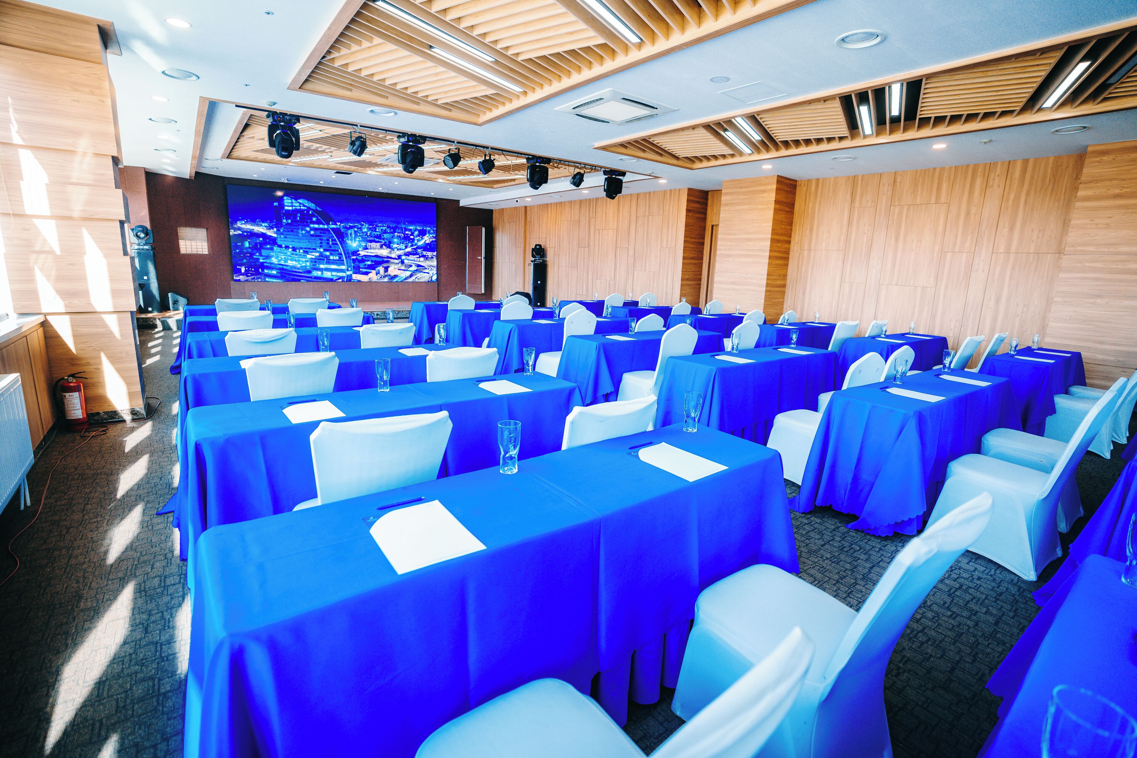 Une salle de conférence avec des rangées de tables recouvertes de nappes bleues et des chaises blanches, un grand écran affichant une vue panoramique de la ville, et des panneaux de bois sur les murs.