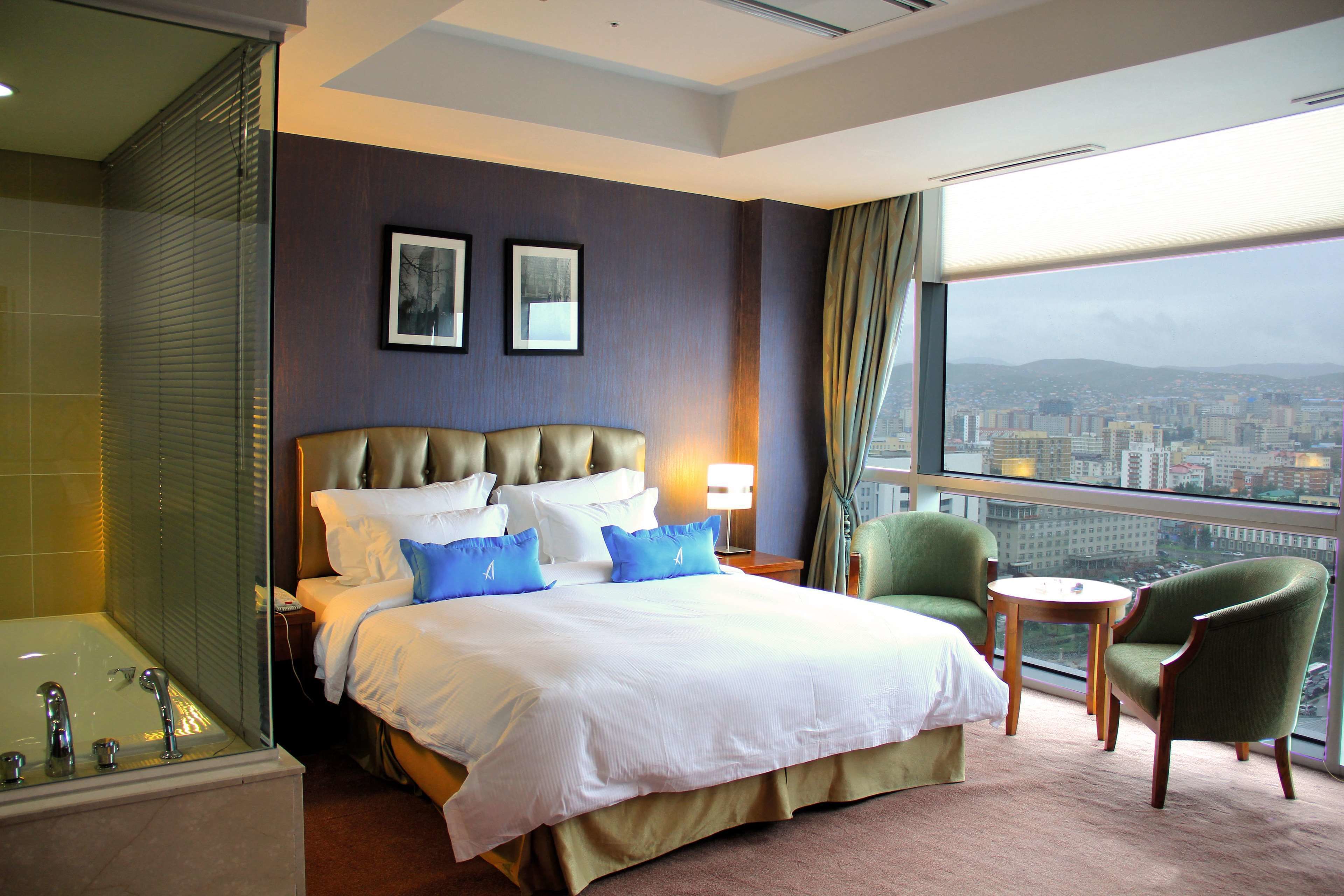 Une chambre d’hôtel avec un grand lit, un coin salon comprenant un fauteuil et une table, une baignoire et une vue sur la ville par la fenêtre.