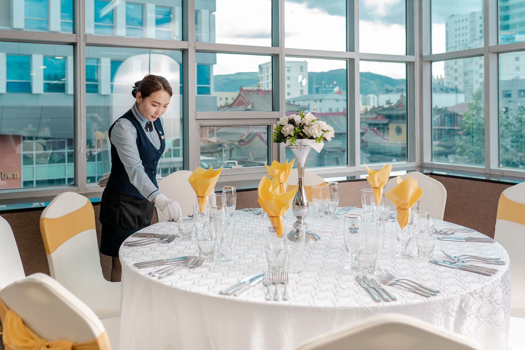 A hotel room with a round dining table set for a meal, large windows offering a view of buildings and hills, and a maid preparing the table.