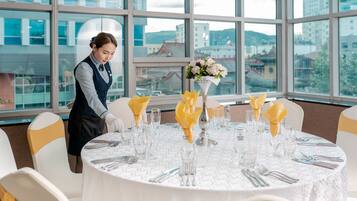 A hotel room with a round dining table set for a meal, large windows offering a view of buildings and hills, and a maid preparing the table.