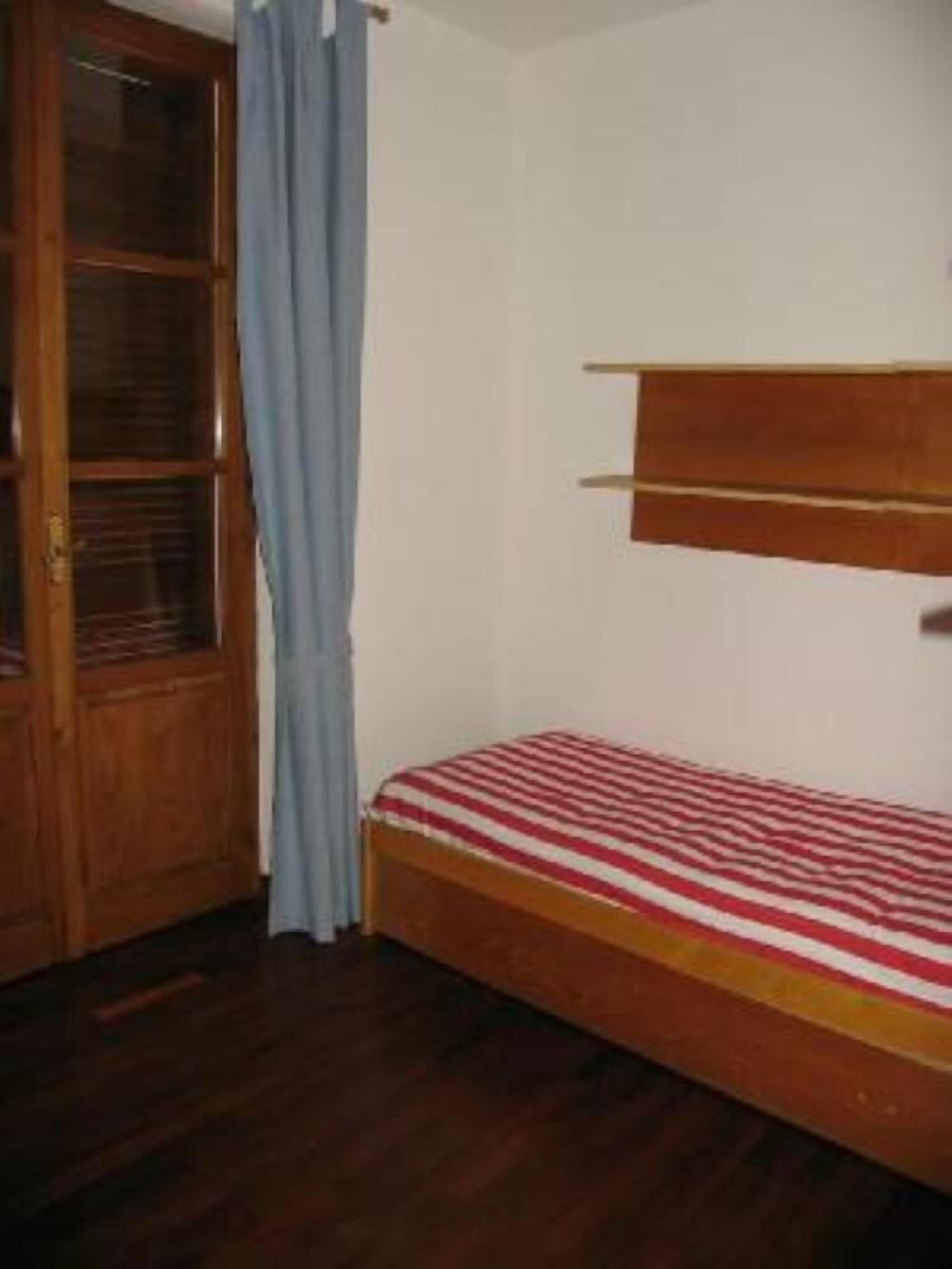 A small, single-bed room with a wooden headboard, a window with curtains, and a wooden wardrobe.