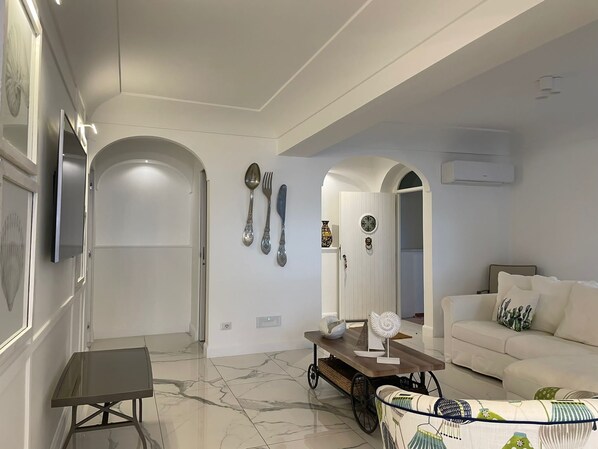 A modern living room with a sofa, a coffee table, and a TV mounted on the wall. There are decorative items on the wall and a small table with a fan. - Villa Arzilla 1 with charming view, w/o steps, parking and private beach (Positano)