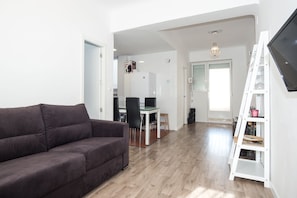 Uma sala de estar moderna com um sofá escuro, uma área de jantar com mesa e cadeiras, e uma estante branca com uma TV na parede.
