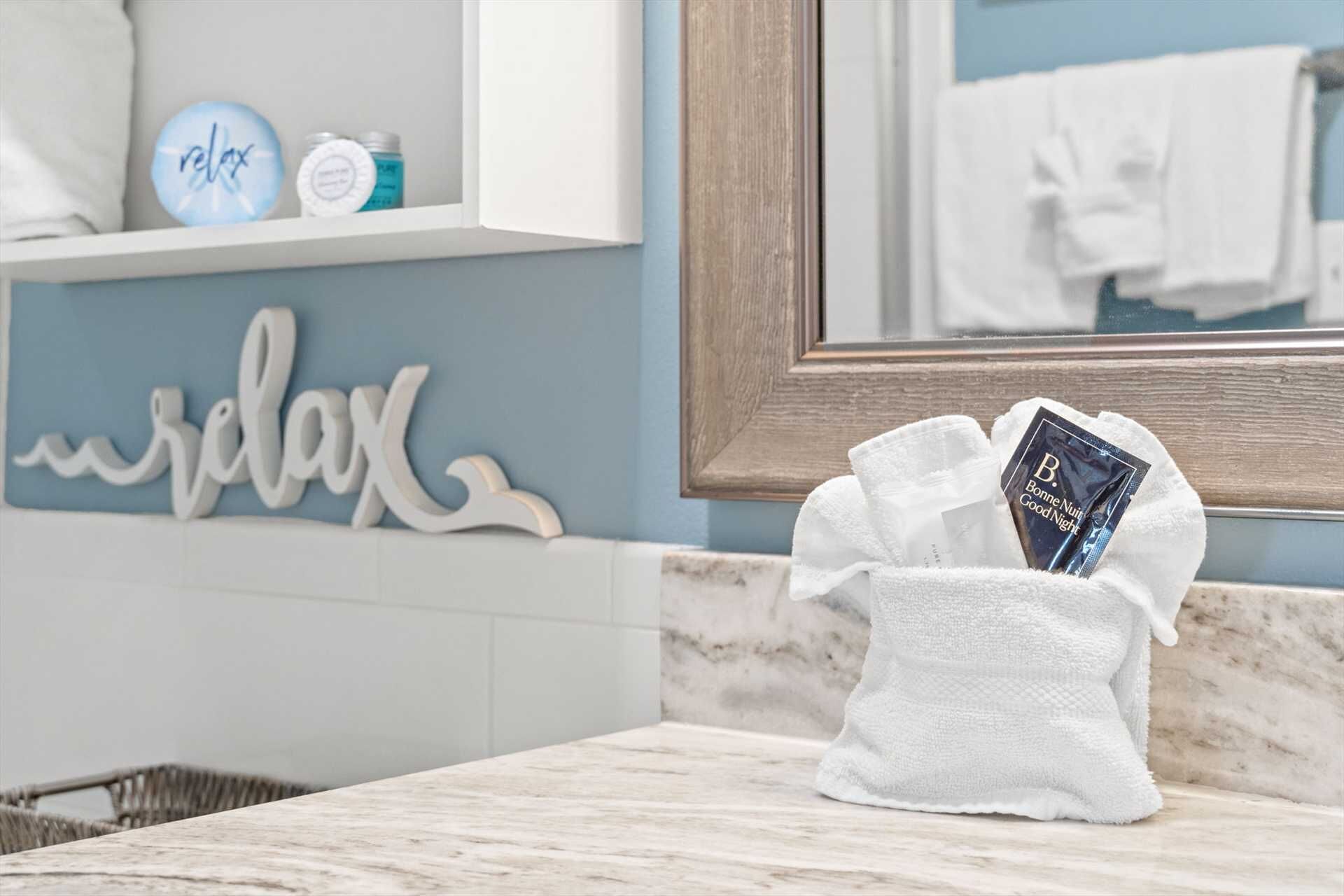 A bathroom with a marble countertop, a towel rack with a white towel, a shelf with a 'relax' sign, and a mirror reflecting a towel.