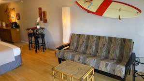 En hyggelig opholdsstue med en stribet sofa, et lille bord, en surfbræt på væggen, en seng og et tekøkken i baggrunden.