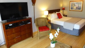 Et hotelværelse med en seng, et fjernsyn på et træskab, en fletstol med rød pude, et natbord med en lampe og et billede i ramme på væggen.