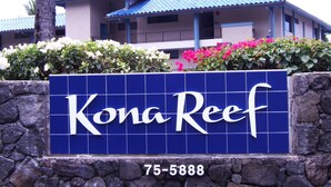 En skylt för Kona Reef, med en stenmur och en byggnad med blå tak i bakgrunden.