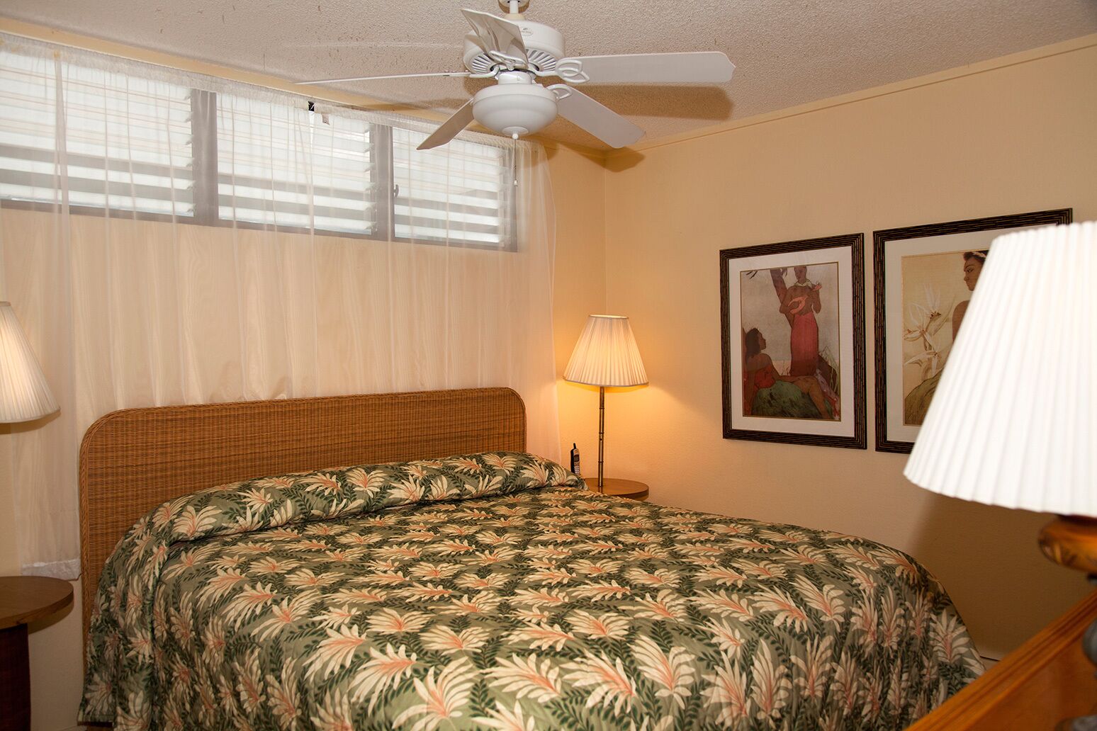 A hotel room with a bed, a ceiling fan, a window with blinds, a bedside lamp, and two framed pictures on the wall.