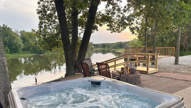 A hot tub with a wooden deck and chairs, surrounded by trees and a lake. - Cedar Bend Cabin with 4 person hot tub!! (Nashua)