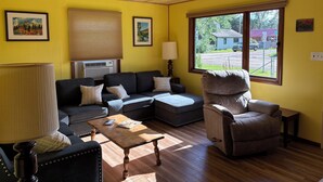 Una habitación con techo de madera, paredes amarillas, un sofá negro, un sillón marrón, una mesa de centro de madera y una ventana con vista a una casa y una calle.