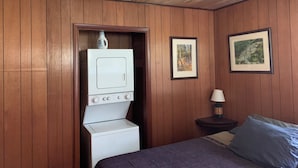 Una habitación con paredes de paneles de madera, una cama con ropa de cama azul, una lavadora blanca y dos cuadros en la pared.