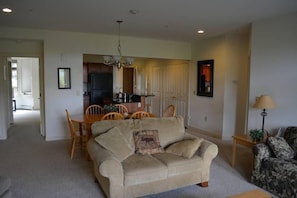A living room with a beige sofa, a dining table with chairs, a chandelier, and a kitchen area in the background.