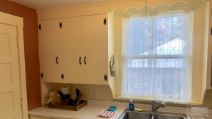 Una cocina con armarios blancos, un ventanal con cortina translúcida, un lavamanos y varios objetos de cocina sobre la encimera.
