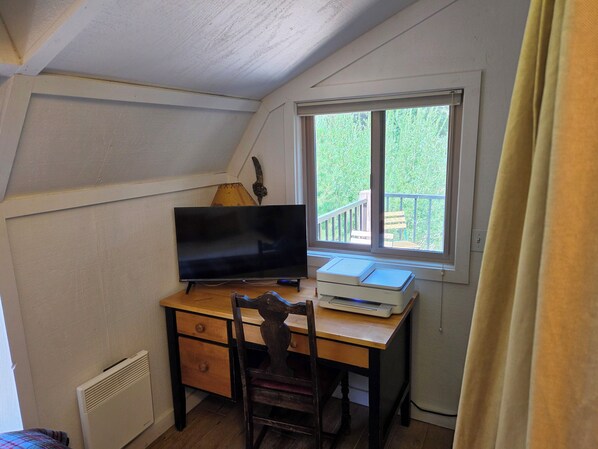 Um quarto com escrivaninha, televisão, cadeira, janela com vista para o verde e radiador.