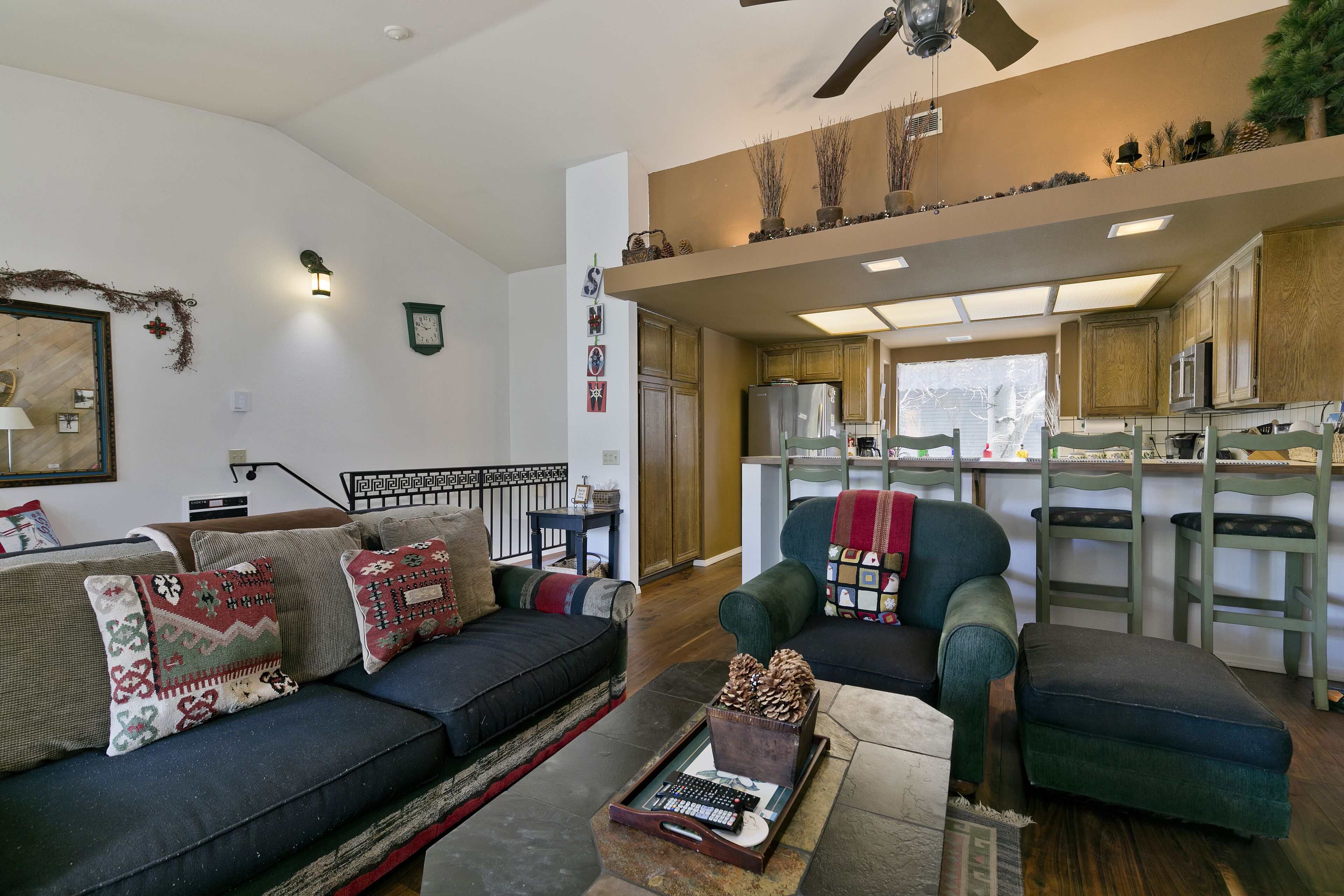 A living room with a sofa, armchairs, and a coffee table. A kitchen is visible in the background with wooden cabinets and a refrigerator.