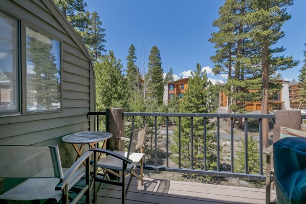 A balcony with a table and chairs, overlooking a forested area with tall trees and a clear sky.