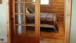 A wooden cabin interior with a bed, a window, and a door with a glass panel. - The North Shore Fisherman's Cabin- historic (Grand Marais)