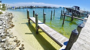 A pier extending into clear water with a rocky shoreline and a distant marina.