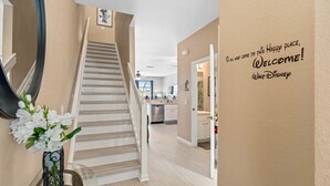 Interior de una casa con una escalera que lleva a una zona de cocina. Un cartel en la pared dice: '¡Bienvenidos! Walt Disney.'