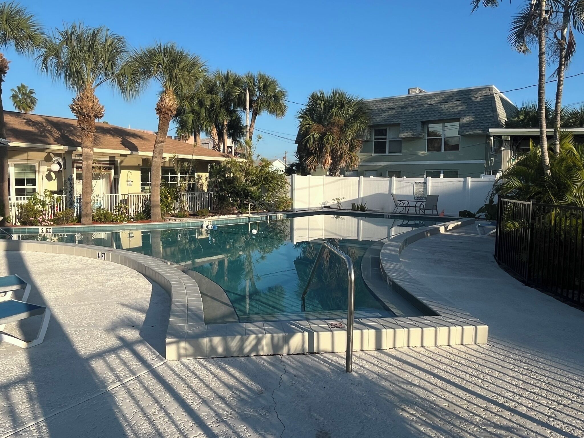 A swimming pool with a tiled deck, surrounded by palm trees and a building with a sloped roof.
