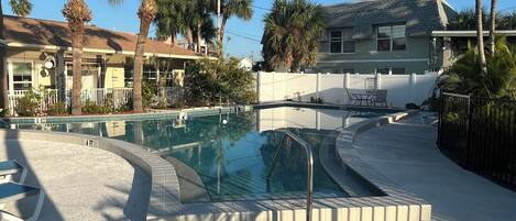 A swimming pool with a tiled deck, surrounded by palm trees and a building with a sloped roof.
