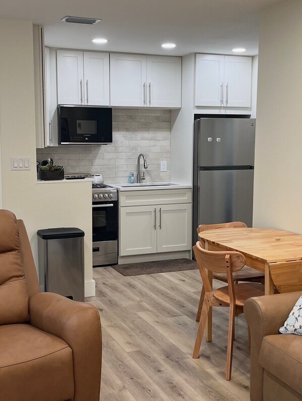 A compact kitchen with white cabinets, a stainless steel refrigerator, a microwave, and a sink. There is a wooden dining table with chairs.
