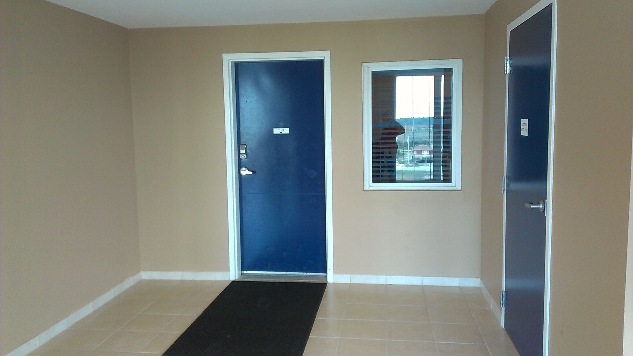 Un pasillo con una puerta azul, una ventana con persianas y un felpudo negro en la entrada.