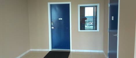 Un pasillo con una puerta azul, una ventana con persianas y un felpudo negro en la entrada.