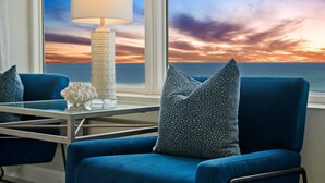 A room with a view of a sunset over the ocean, a blue armchair with patterned cushions, a white lamp, and a small decorative item on a glass table.