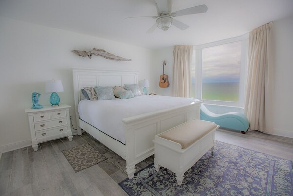 A bedroom with a bed, a nightstand, a bench, a guitar, and a view of the ocean.