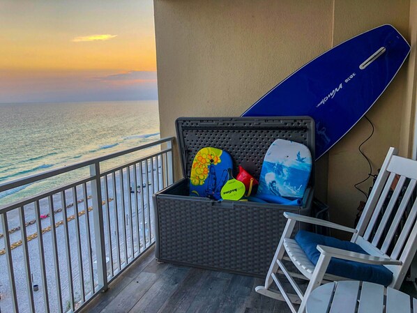 Un balcón con vista al océano, un baúl con tablas de surf y una mecedora blanca.