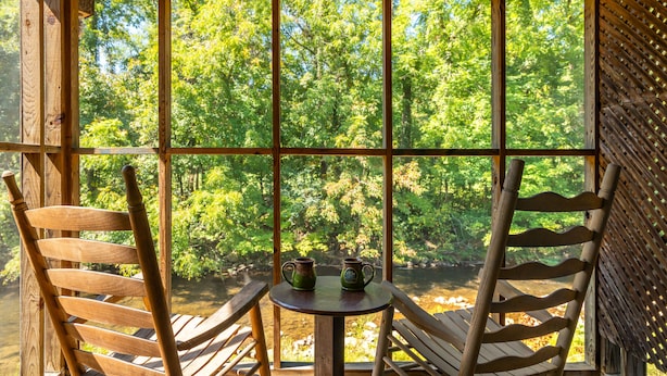 Puinen mökki, jossa on suuri ikkuna, josta avautuu näkymä metsäiselle alueelle. Sisällä on kaksi puista tuolia ja pieni pöytä, jonka päällä on kaksi vihreää mukea.