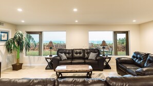 A spacious living room with a leather sofa, a coffee table, and a large window offering a view of the outdoors. - Private Oceanfront Estate • Pickleball court • Pool • Spa • Golden Sands® Pearl (Indialantic)