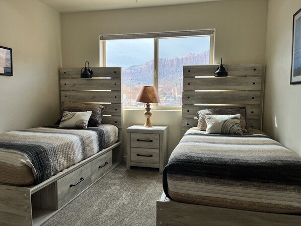 A bedroom with two beds, a nightstand with a lamp, and a window with a view of mountains.