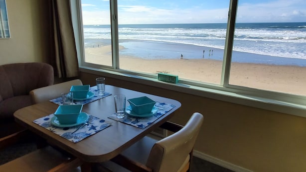 Un coin repas avec une table dressée pour deux personnes, une fenêtre donnant sur la plage et un panneau indiquant le nom de la plage.