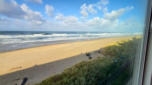 Une vue sur la plage depuis une fenêtre, incluant l’océan, la plage de sable et les dunes herbeuses.