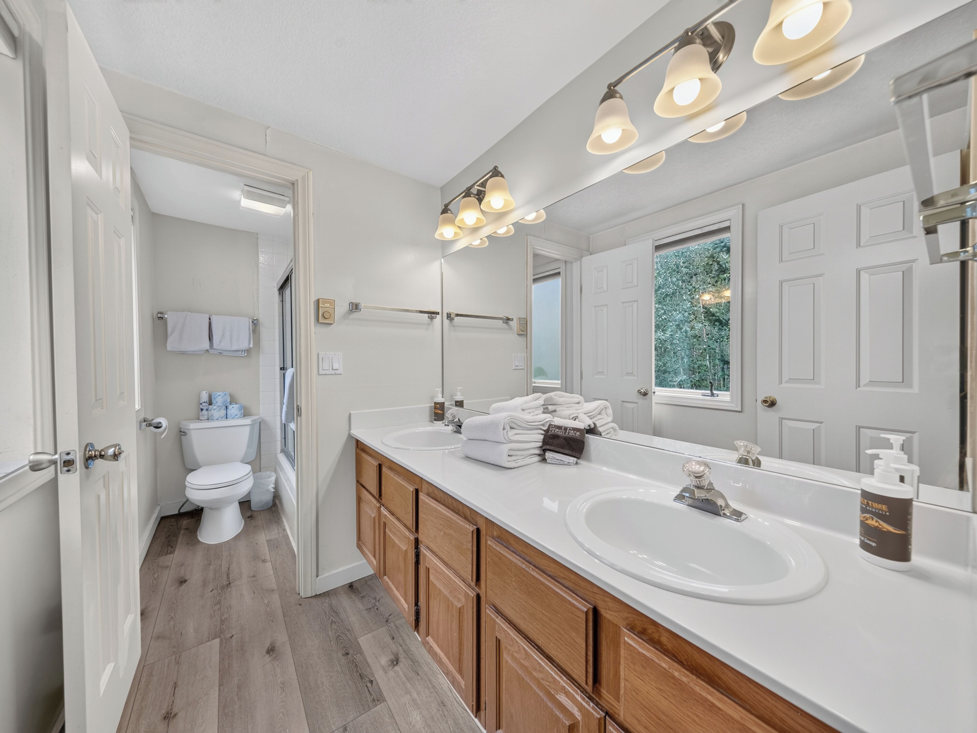 Une salle de bain avec un lavabo blanc, des meubles en bois, un grand miroir et une fenêtre donnant sur des arbres.