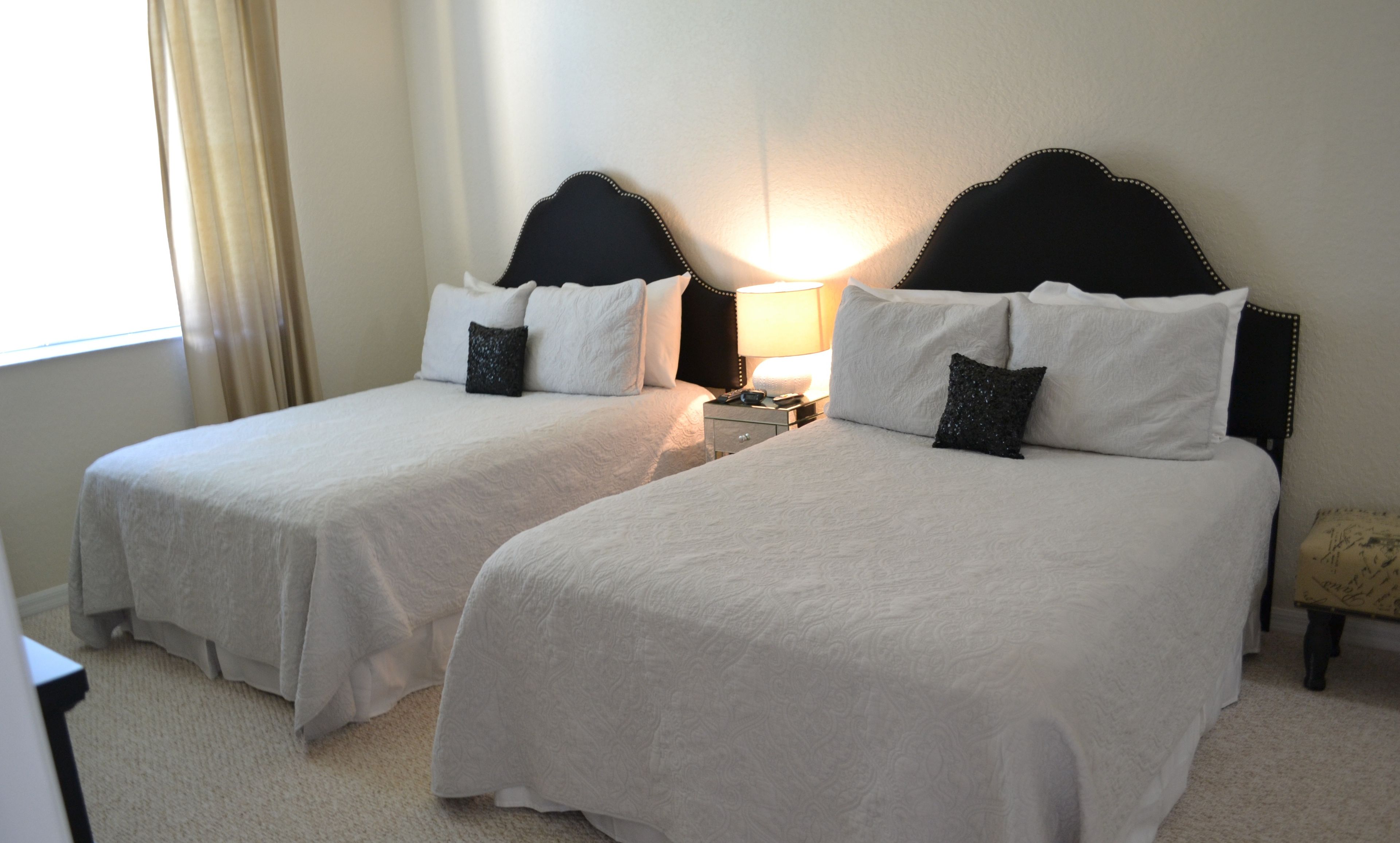 Un dormitorio de invitados con dos camas, cada una con ropa de cama blanca y almohadas decorativas, una mesita de noche con lámpara y una ventana con cortinas.