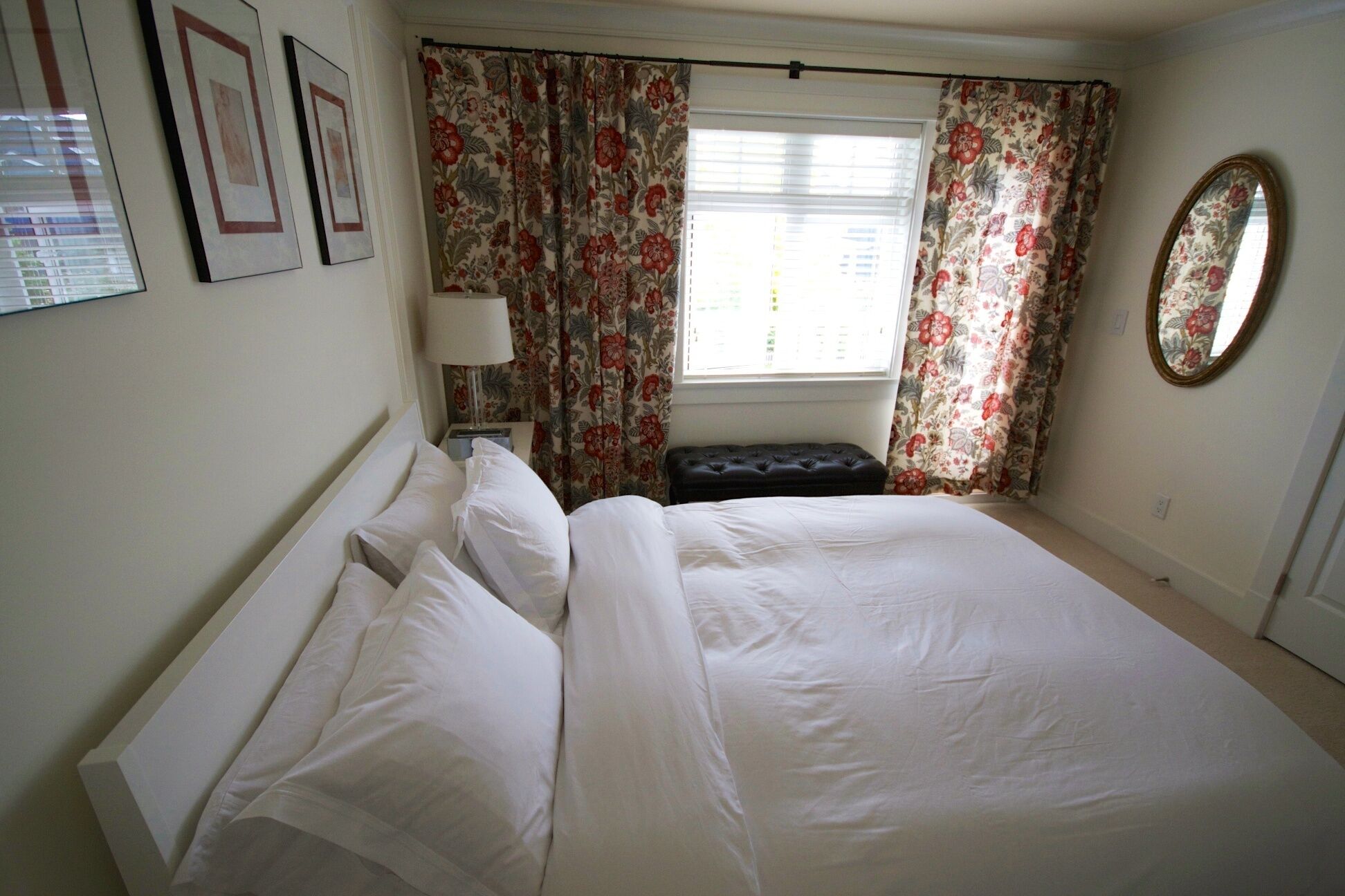 Un lit bien fait, avec du linge de lit blanc, une lampe de chevet, un rideau à motifs floraux et un miroir rond accroché au mur.