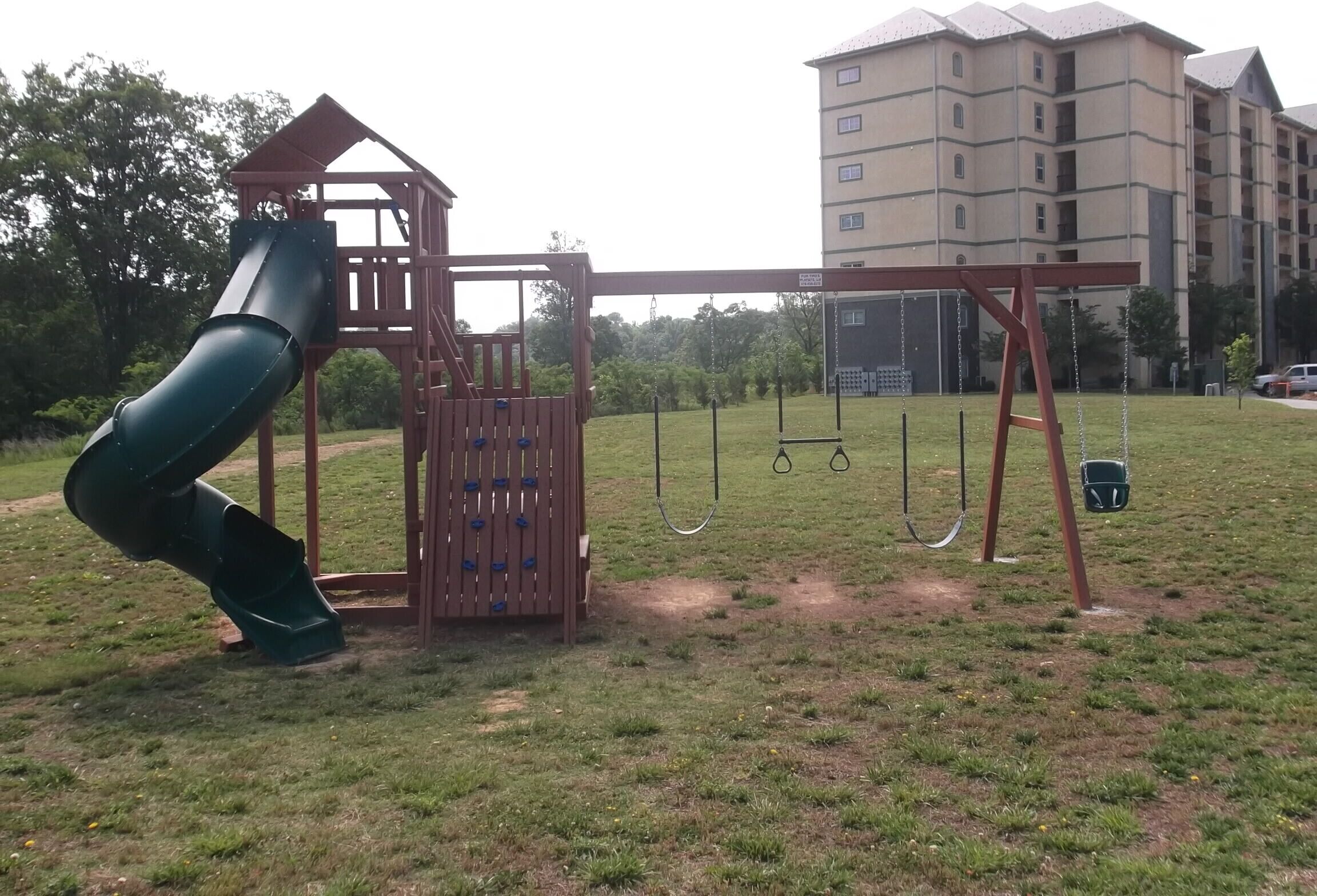 A playground with a slide and swings, surrounded by grass and a building in the background.