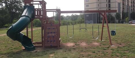 A playground with a slide and swings, surrounded by grass and a building in the background.