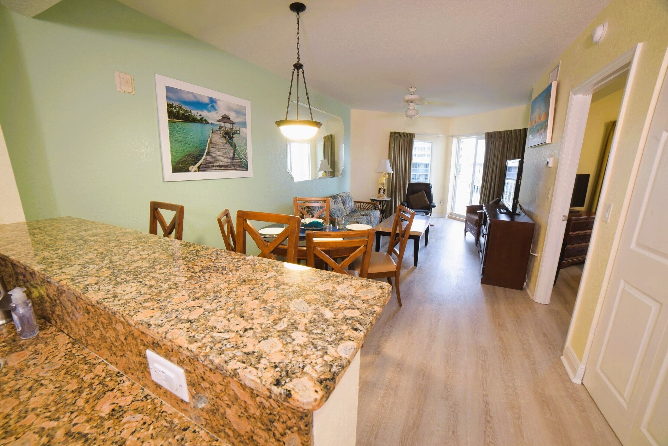 A kitchen with granite countertops, wooden chairs, and a dining table. There is a hallway leading to a living area with a couch and a TV.