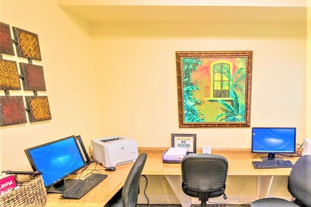A workspace with two computer monitors, a printer, and a desk with a chair. There is a framed painting on the wall and a sign that says "PLEASE NO FOOD OR DRINKS".
